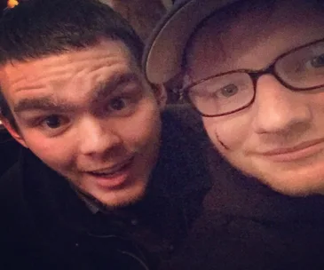 Two individuals taking a selfie; one has a noticeable mark on his cheek, wearing glasses and a cap.