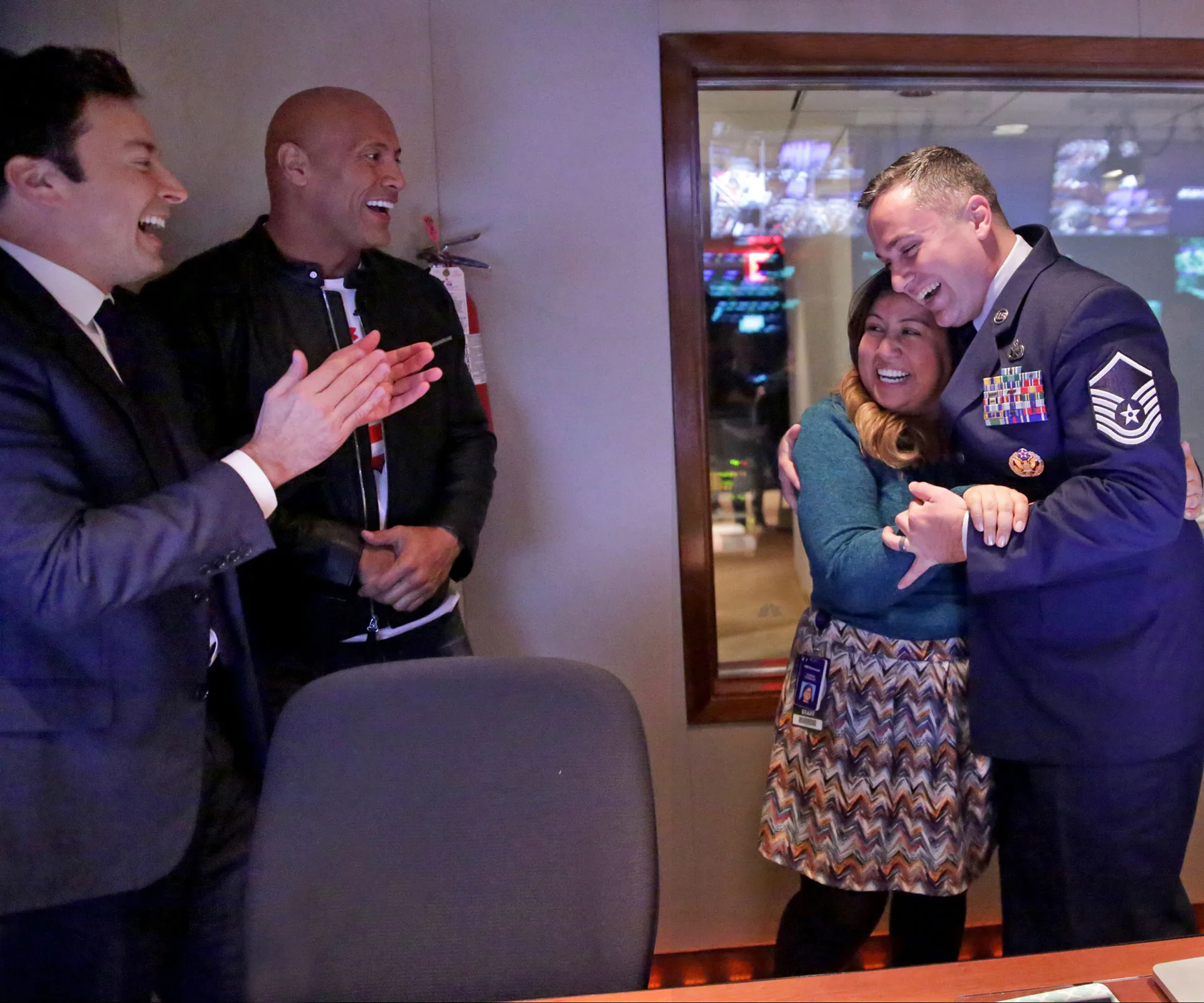 Group celebrating a joyful reunion; two men clapping, while a military officer hugs a smiling woman in a control room.