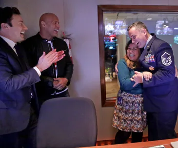 Group celebrating a joyful reunion; two men clapping, while a military officer hugs a smiling woman in a control room.