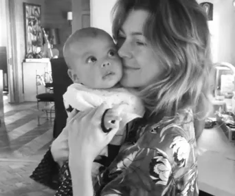 A woman smiles while holding a baby in her arms, looking at the baby lovingly in a cozy home setting.