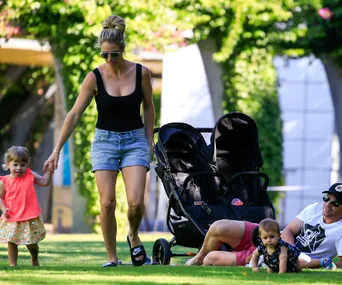 Family enjoys a sunny day in the park, mother walks with daughter, father relaxes near stroller with baby nearby.
