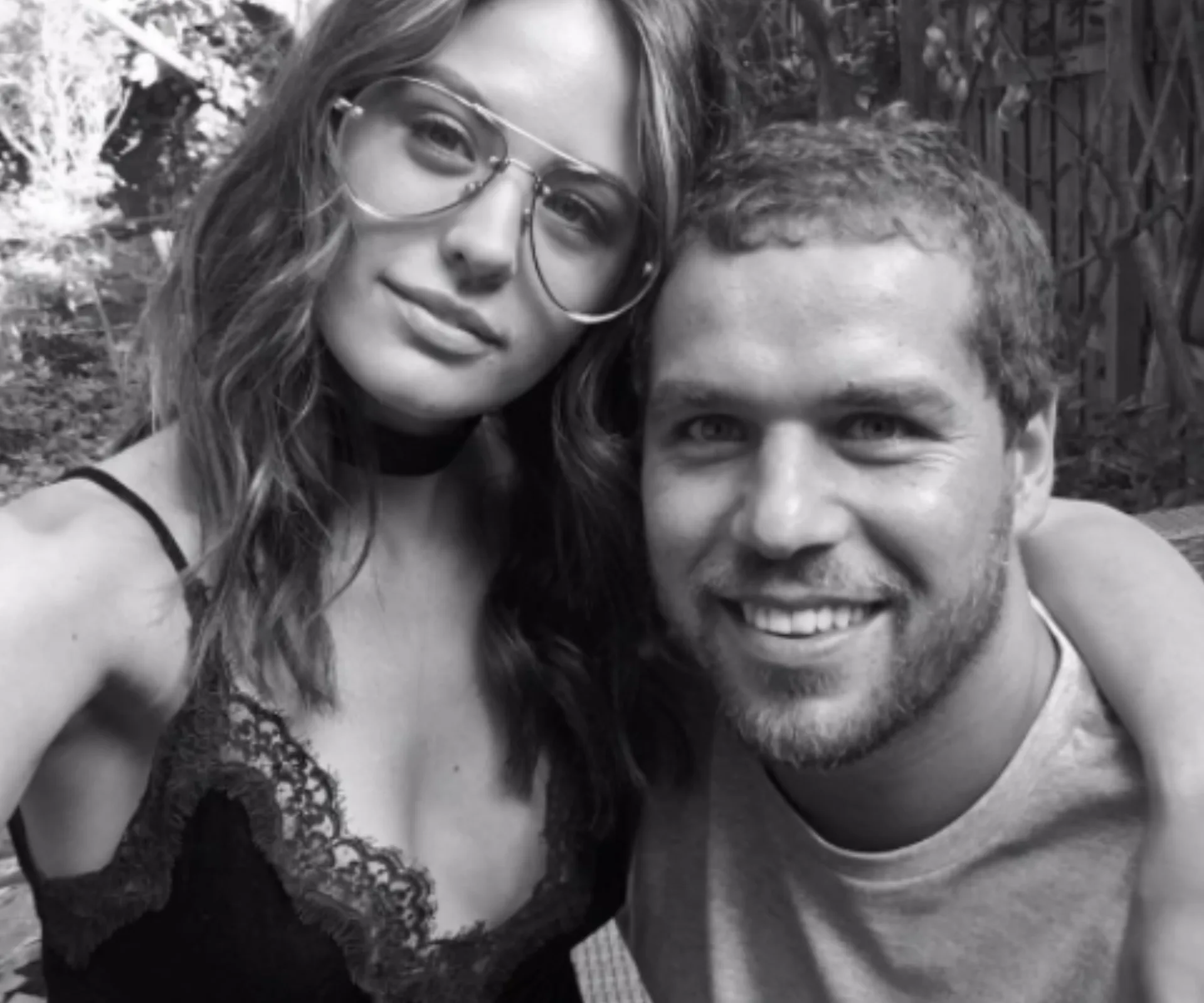 Two people pose for a selfie outdoors, smiling. The woman wears glasses and a lace-trimmed top; the man has short hair and a beard.