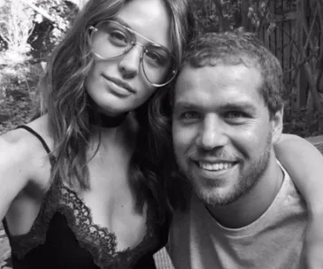 Two people pose for a selfie outdoors, smiling. The woman wears glasses and a lace-trimmed top; the man has short hair and a beard.