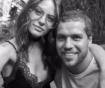 Two people pose for a selfie outdoors, smiling. The woman wears glasses and a lace-trimmed top; the man has short hair and a beard.