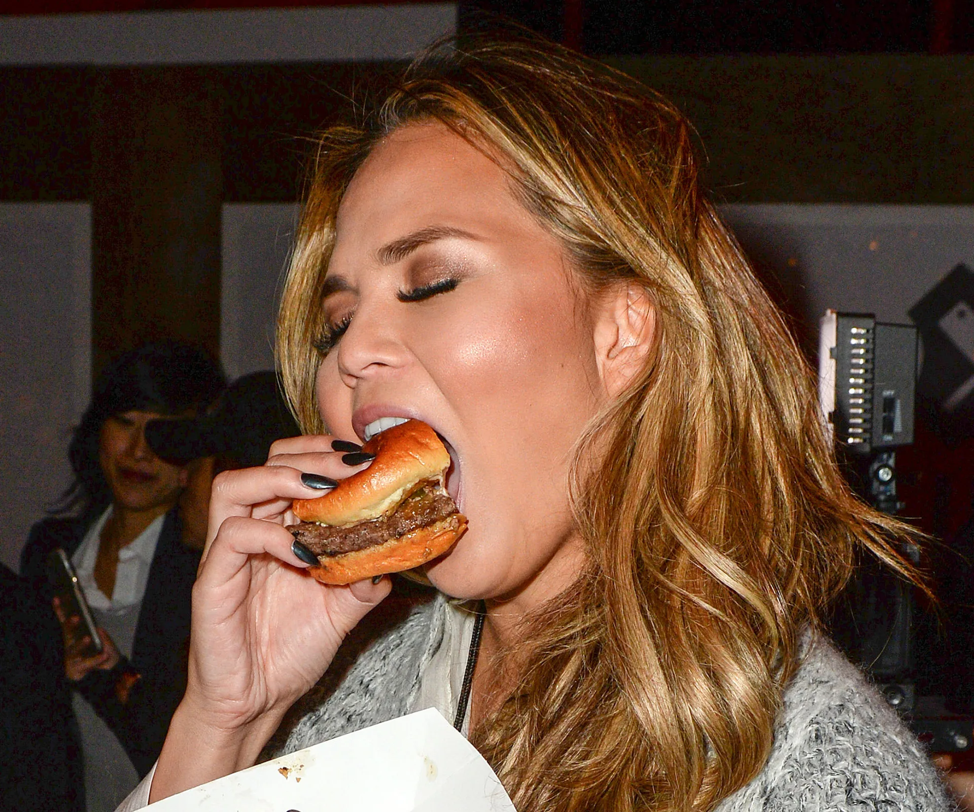 A person is taking a bite from a cheeseburger at an indoor event with people in the background.