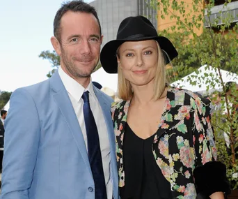 A couple smiling outdoors; she wears a floral blazer and black hat, he is in a light blue suit.