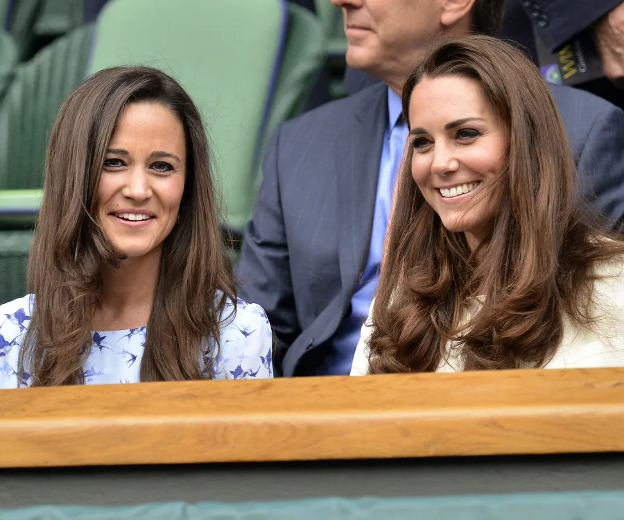 Two women sitting in a stadium, both smiling and wearing elegant attire, with people seated behind them.