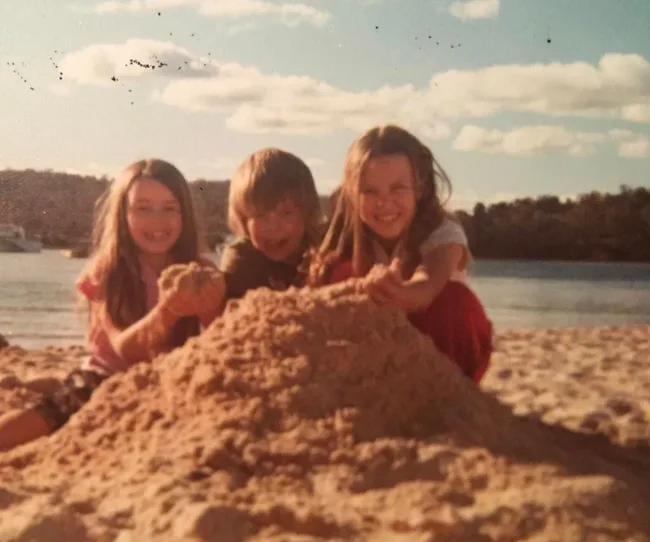 Three children smiling while building a sandcastle on a sunny beach day.