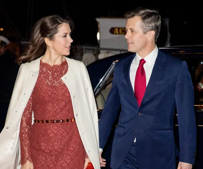 Couple in formal attire arriving at an event, woman in lace dress with coat, man in suit with red tie.