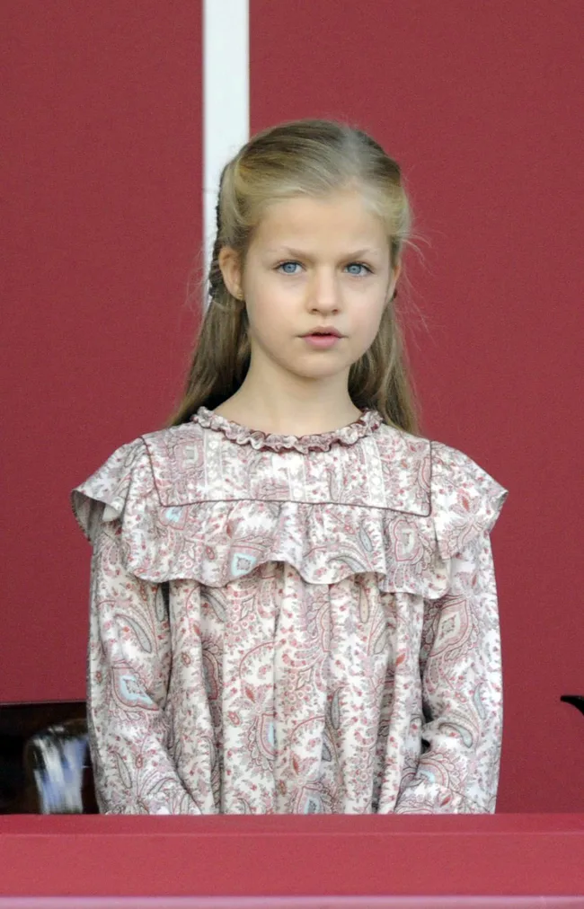 A young girl with long blonde hair in a patterned dress against a red background.
