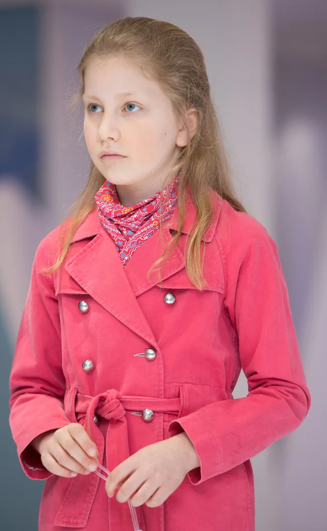 Young girl in a pink coat and scarf, standing indoors, looking thoughtful.