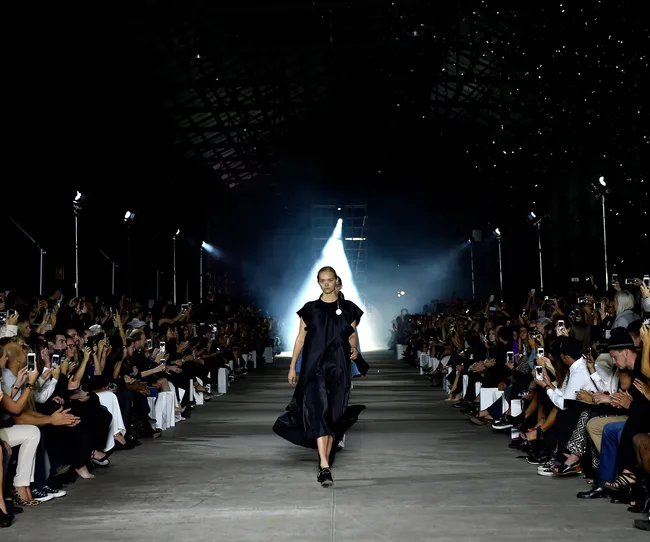 Model walking down a catwalk in a dark dress during a fashion show, with an audience taking photos on both sides.