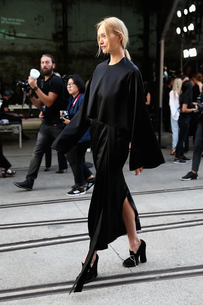 Model in a flowing black dress walks on a runway, photographers in the background capturing the moment.