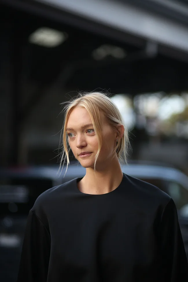A woman with blonde hair, wearing a black top, stands outdoors with a thoughtful expression.