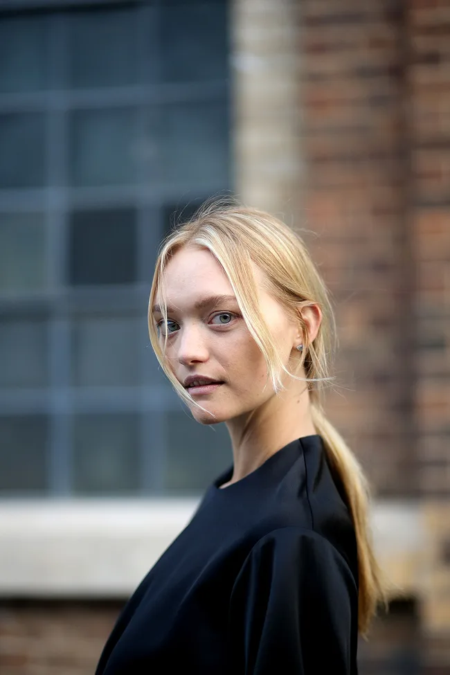 Blonde woman with tied hair and black attire, gazing over shoulder, soft focus background of a brick building and window.