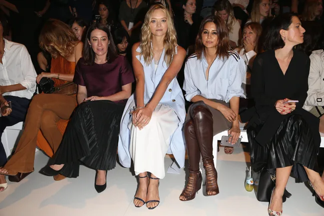 Front row at fashion show, six women seated, one in light blue dress, one in maroon top, others in varied outfits, audience behind.
