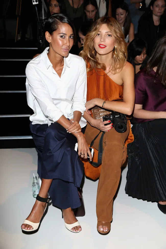 Two women seated at a fashion event; one in a white shirt, the other in an orange top holding a camera.