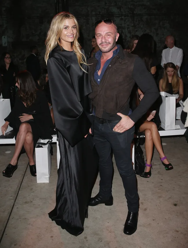 Two people posing together in stylish outfits at a fashion event, with seated audience in the background.