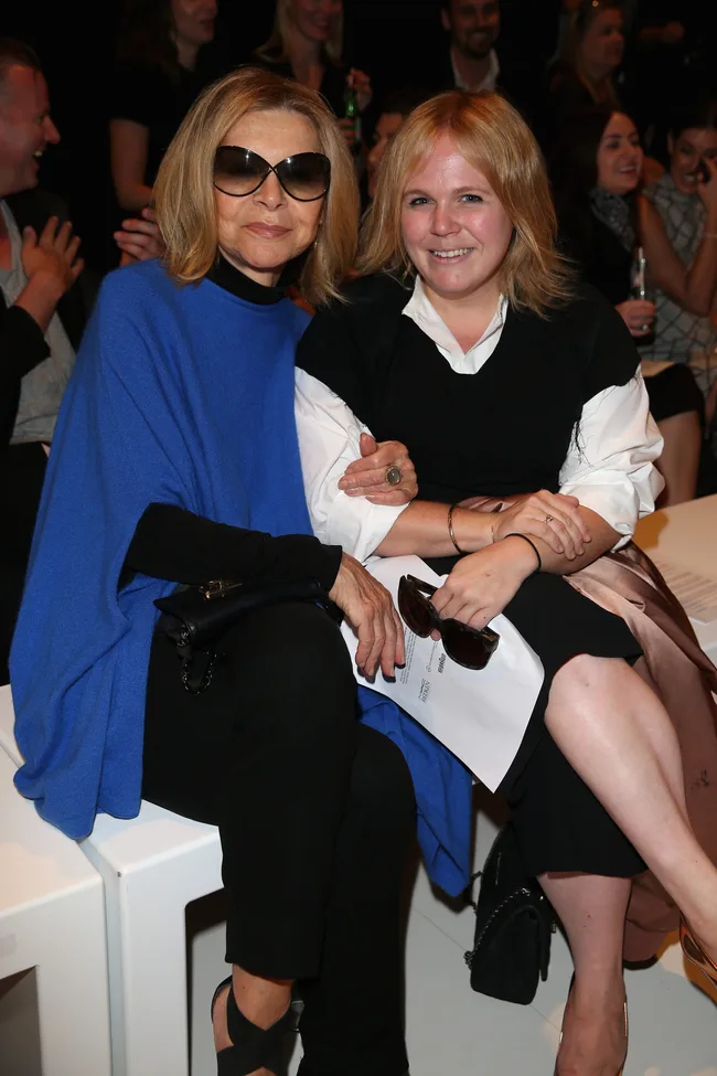 Two women seated, one in a blue top with sunglasses, the other in black and white, smiling at an event.