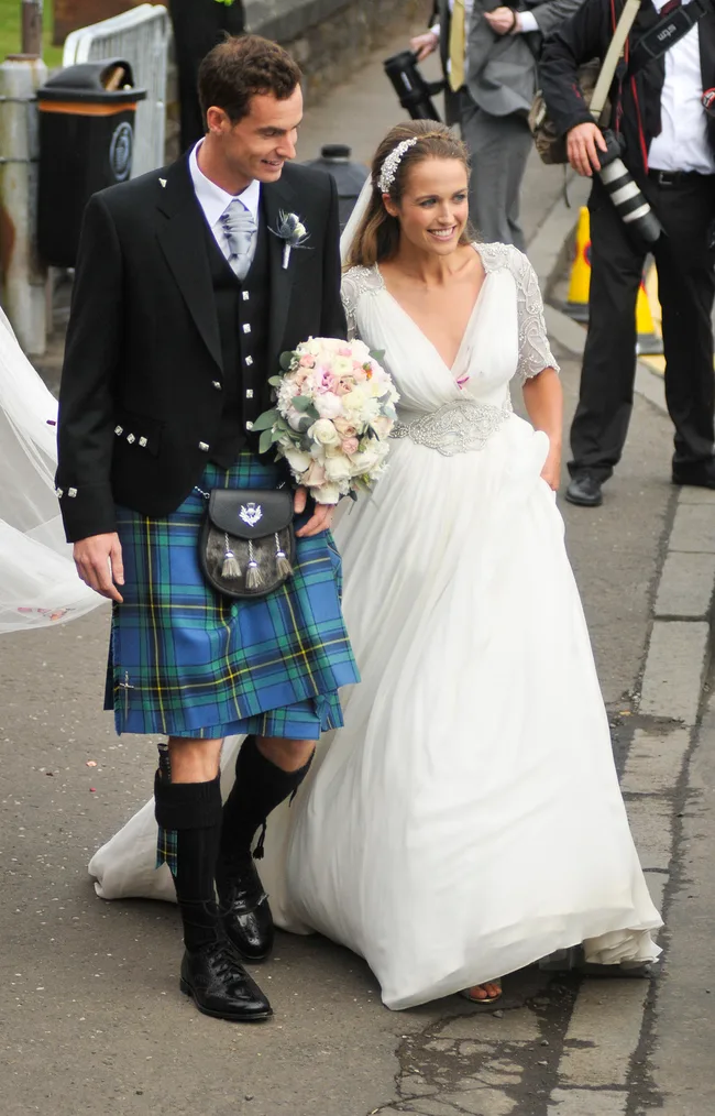 Bride in white gown and groom in a kilt, walking and smiling, with photographers in the background.