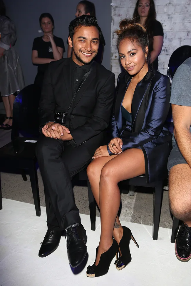 Two people seated, smiling, dressed in stylish evening wear at an event, with others visible in the background.