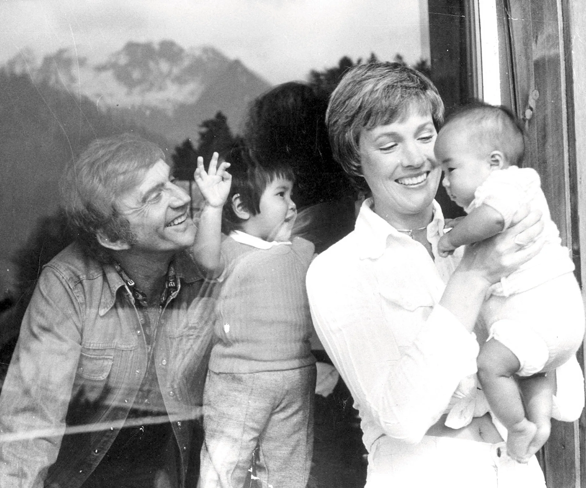 Lady holding a baby with a smiling man and child visible through glass; background shows mountains.