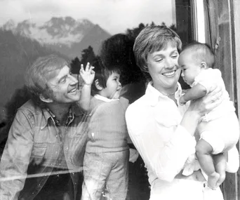 Lady holding a baby with a smiling man and child visible through glass; background shows mountains.