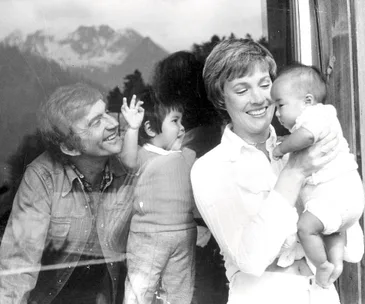 Lady holding a baby with a smiling man and child visible through glass; background shows mountains.