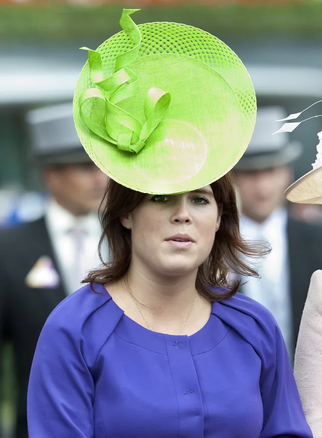 Woman in blue dress wearing a large green circular hat at an outdoor event with people in background.