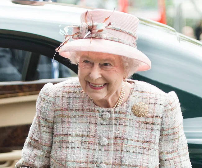 An elderly woman in a pink hat and tweed jacket, smiling near a car.