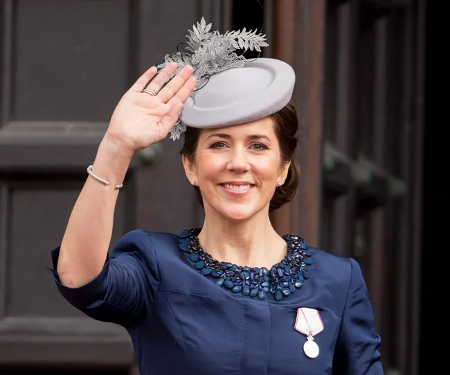 A woman in a blue outfit and decorative gray hat smiling and waving outdoors.