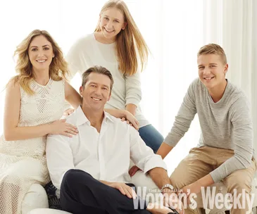 A family of four smiling, including a man, woman, girl, and boy, posing together in light clothing.