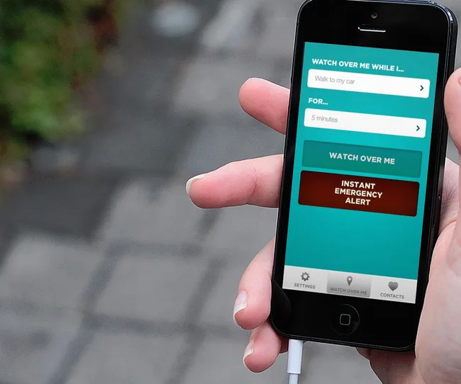 Hand holds smartphone displaying a safety app with options like "Watch Over Me" and "Instant Emergency Alert."