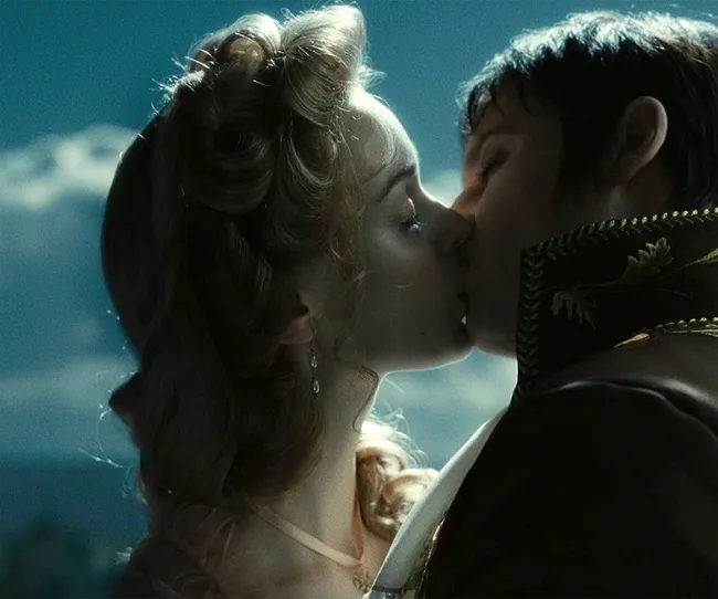 A man and woman, dressed in period costumes, kiss under the night sky.