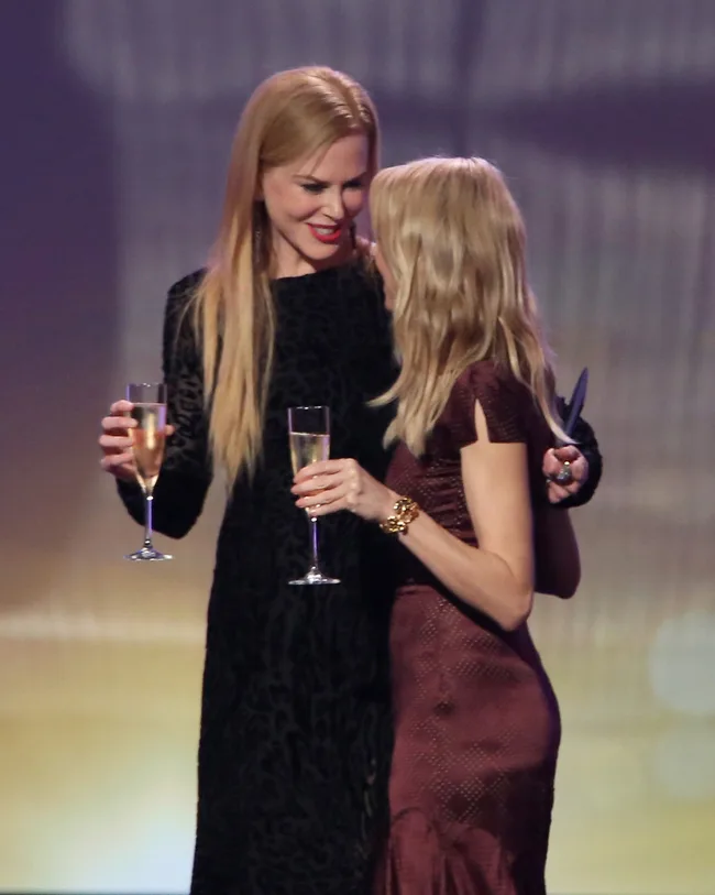 Two women in elegant dresses embrace, holding champagne glasses, on a stage with soft lighting in the background.