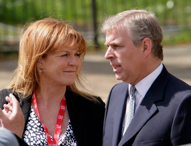 Two people are talking outdoors; the woman has red hair and is wearing a patterned dress, the man is in a suit and tie.