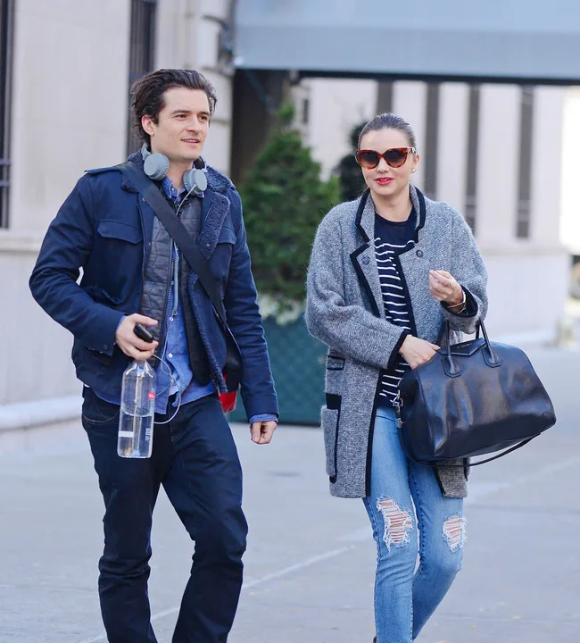 Two people walking on a sidewalk; one holding a water bottle and wearing headphones, the other in sunglasses with a handbag.