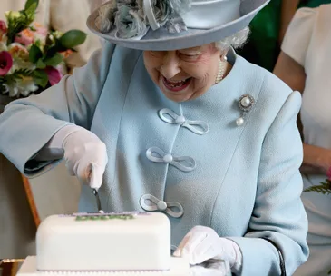 Elderly woman in blue suit and hat, smiling as she cuts a white cake with flowers in the background.