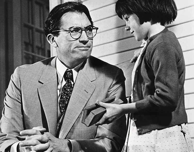 Man in a suit sits, listening to a young girl holding a book, on a porch.