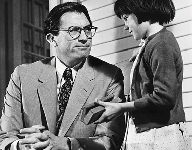 Man in a suit sits, listening to a young girl holding a book, on a porch. 