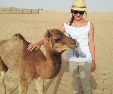 Woman in sunglasses and hat smiling, petting a camel calf in a desert landscape.