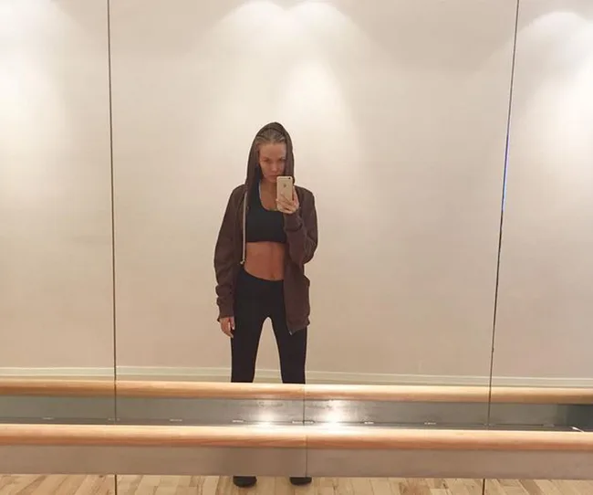 Person taking a selfie in a mirror, wearing a hoodie and workout attire in a dance studio.