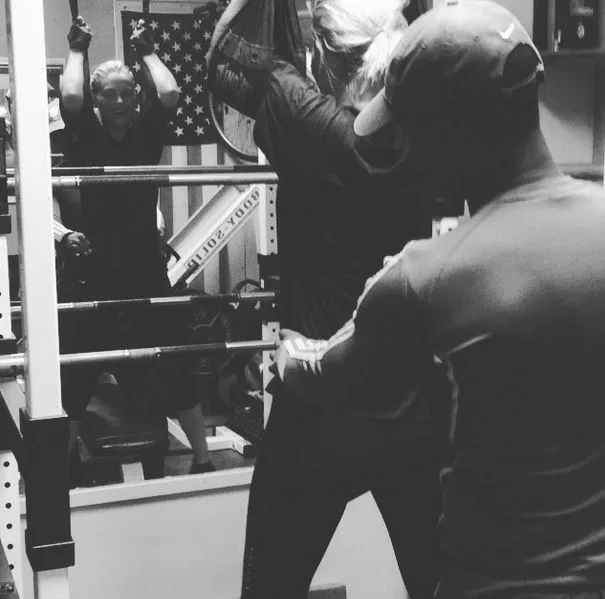 Gym workout with a trainer assisting a person lifting weights in front of a mirror; American flag visible.