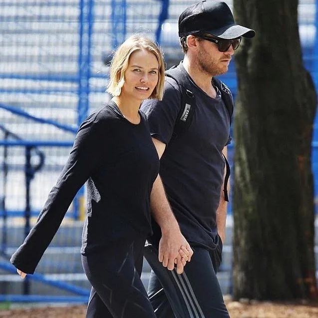 Man and woman walking hand in hand outdoors, both wearing black casual attire; background features blue fencing.