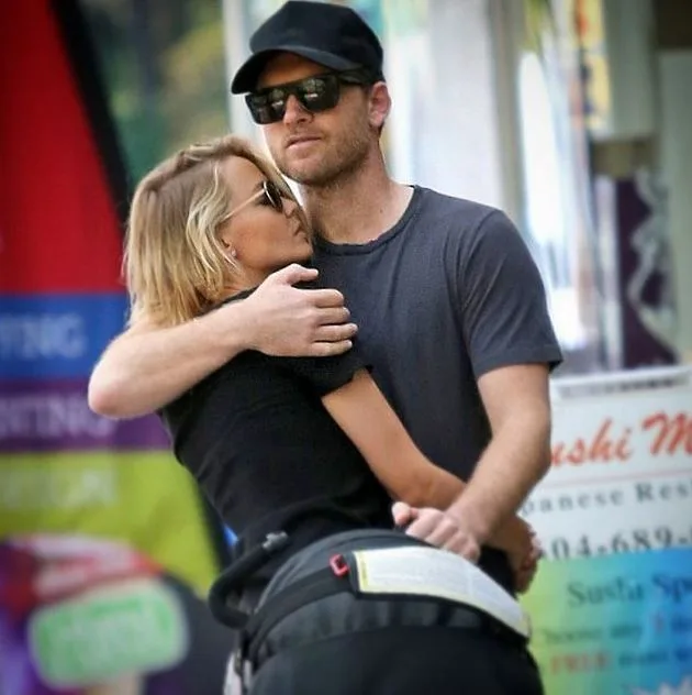 Couple embraces while standing beside a stroller, outdoors, both wearing sunglasses and casual clothes.