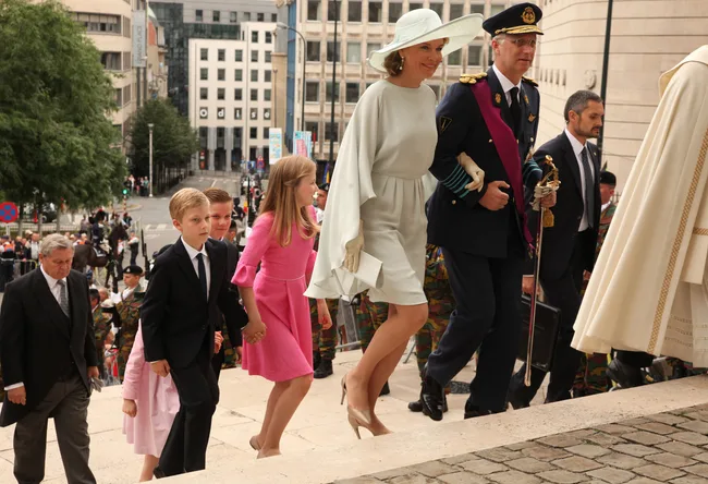 A group of formally dressed people, including a woman in a hat, ascend steps outside a building.