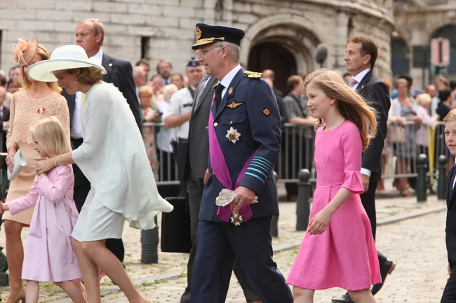 Royal family walking; man in military uniform, woman in white, children in pink dresses on cobbled street, crowd behind.