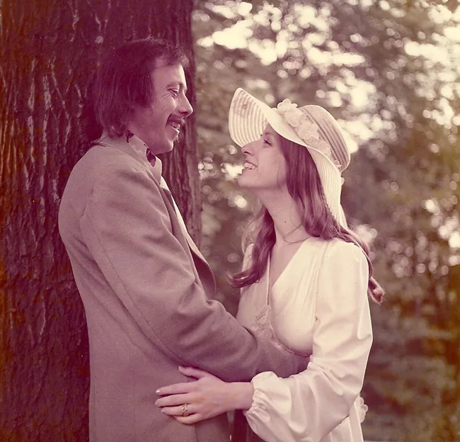 A couple in vintage attire smiles affectionately near a tree, with the woman wearing a wide-brimmed hat.
