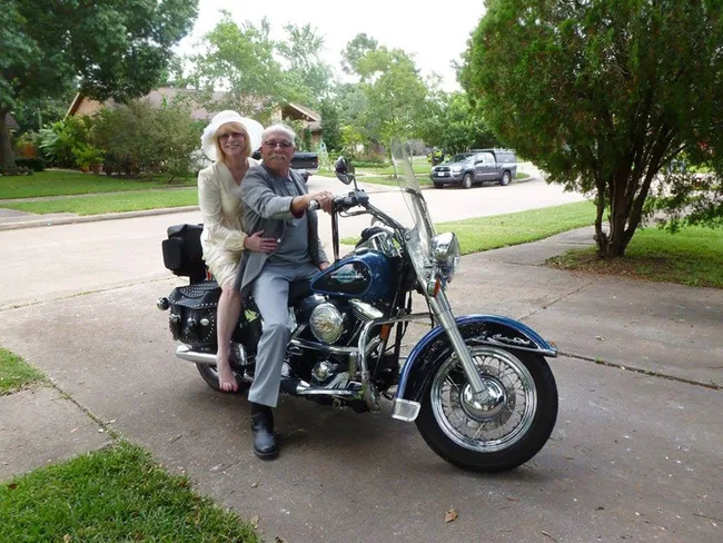 A couple sits on a Harley-Davidson motorcycle in a suburban neighborhood driveway, surrounded by greenery.