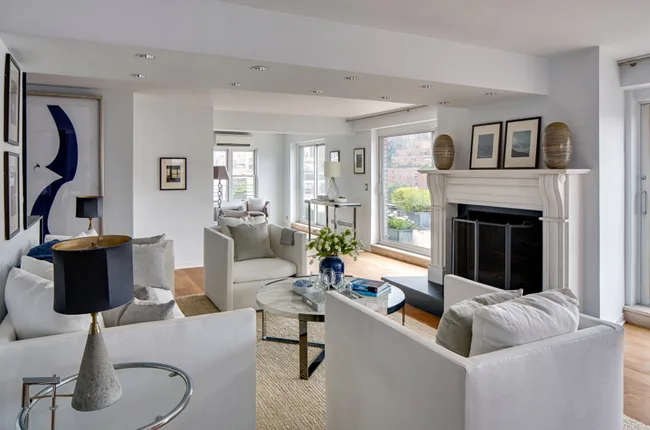 Modern living room with white sofas, a fireplace, framed art, large windows, and a round coffee table with flowers.